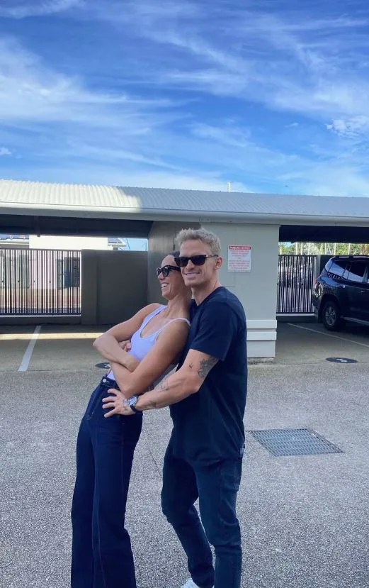 A man and woman in sunglasses pose playfully in a parking lot under a blue sky.