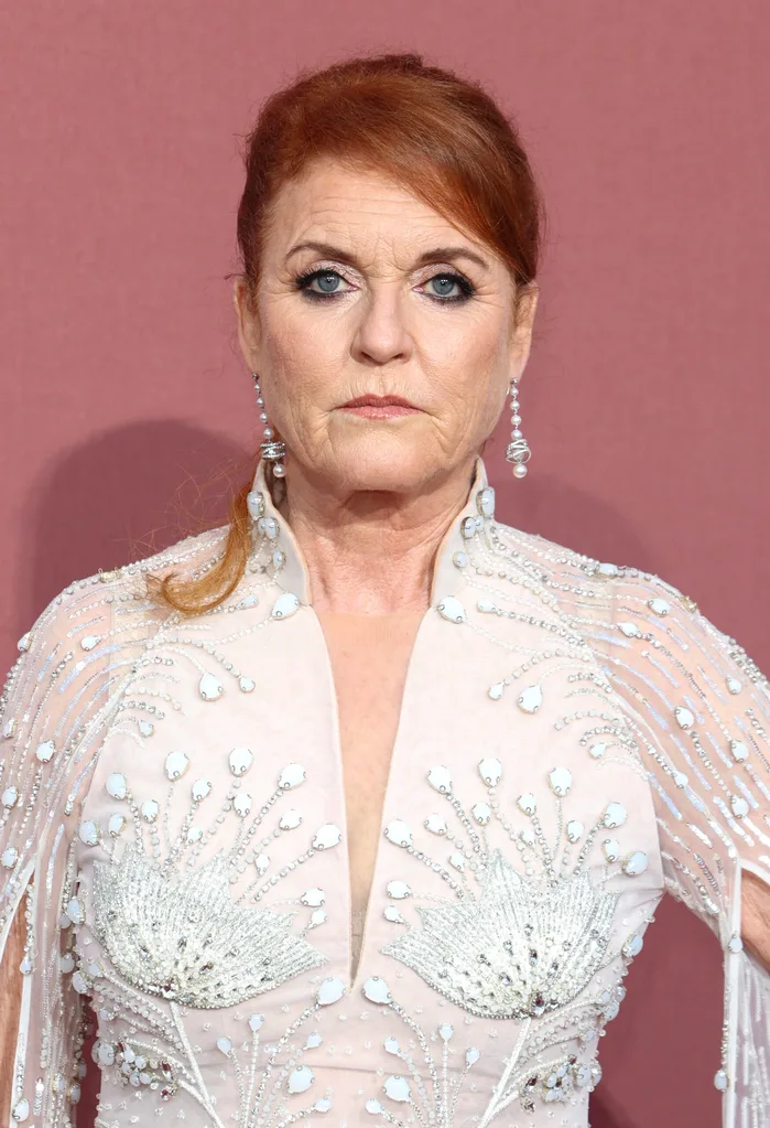 A woman with red hair in an ornate white gown with floral patterns, standing against a maroon background.