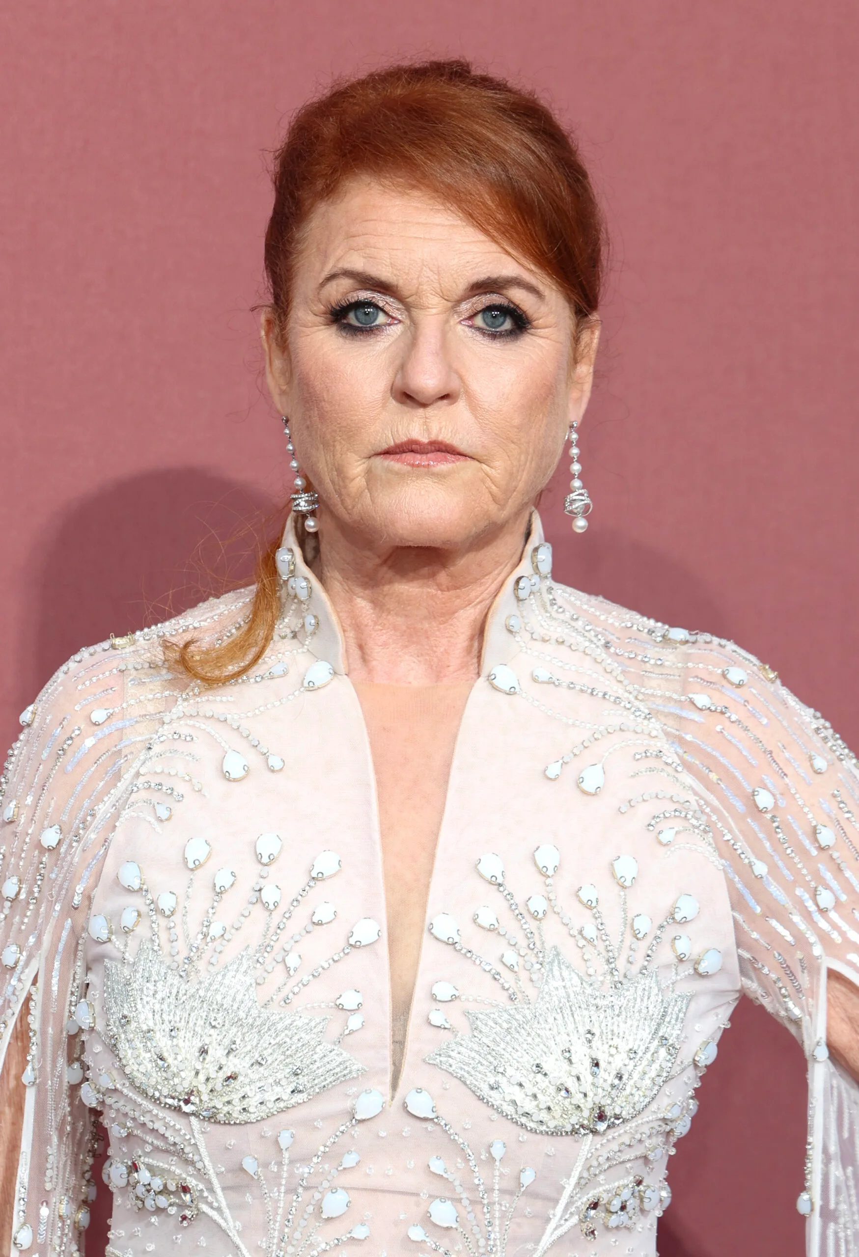 A woman with red hair in an ornate white gown with floral patterns, standing against a maroon background.