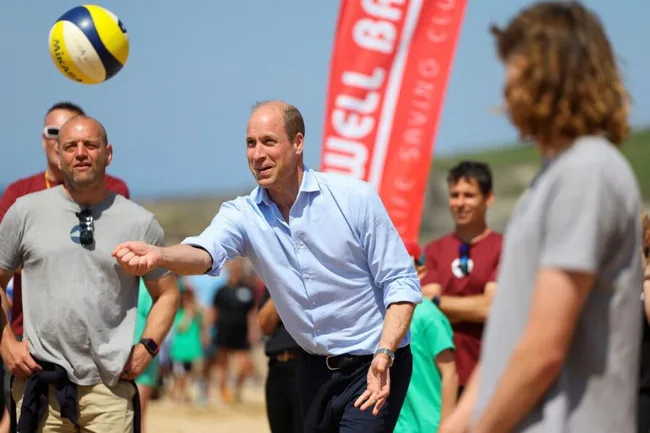 Prince playing volleyball on a sunny beach with spectators in the background.