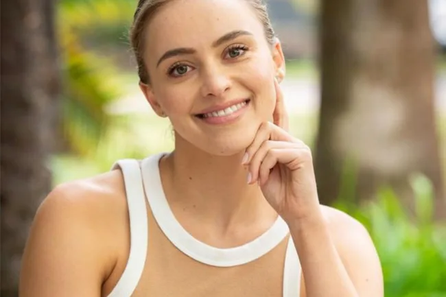 Smiling woman in a sleeveless top, sitting outdoors with a blurred nature background.