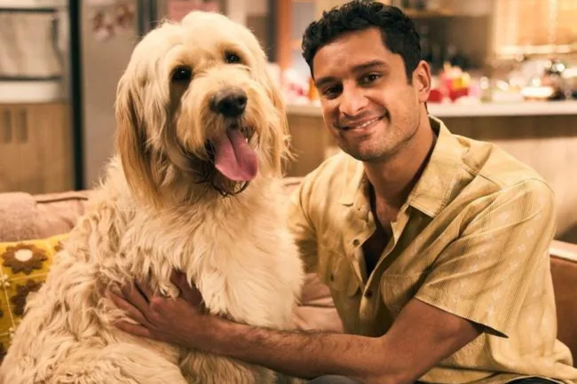 A man smiling beside a large fluffy dog on a couch, in a warmly lit room.