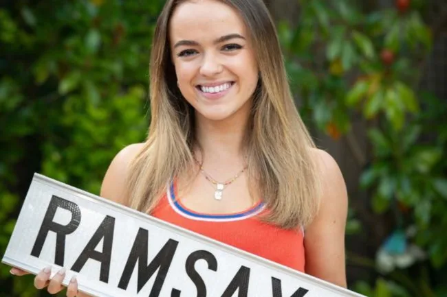 Young woman smiling, holding a sign with "RAMSAY" in bold letters, green foliage in the background.