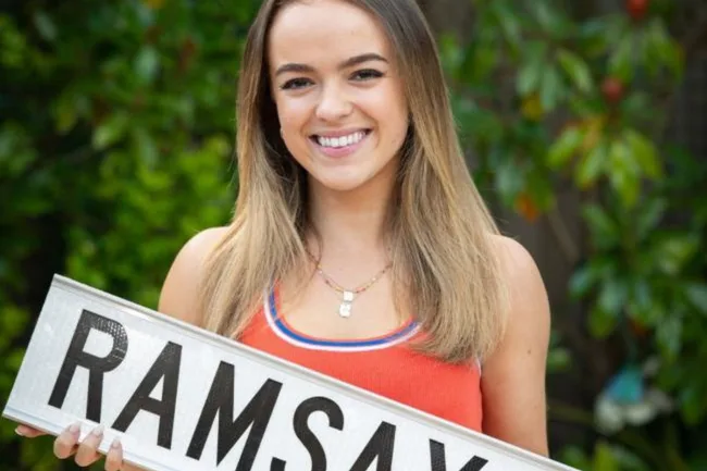 Young woman smiling, holding a sign with "RAMSAY" in bold letters, green foliage in the background.