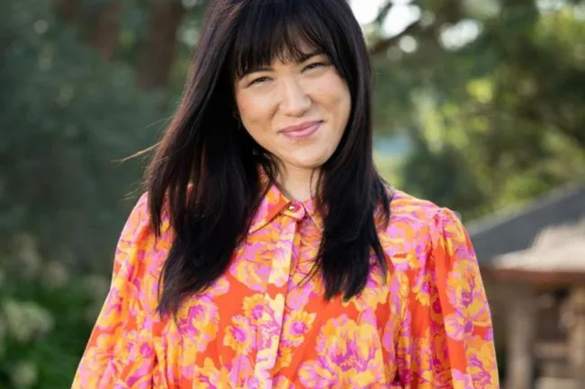A woman with long dark hair smiles, wearing a bright floral blouse, outdoors with trees in the background.