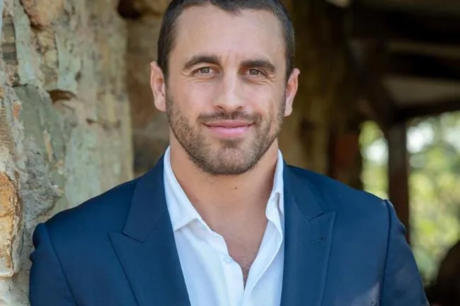 A man in a blue suit and white shirt stands in front of a stone wall, smiling warmly.