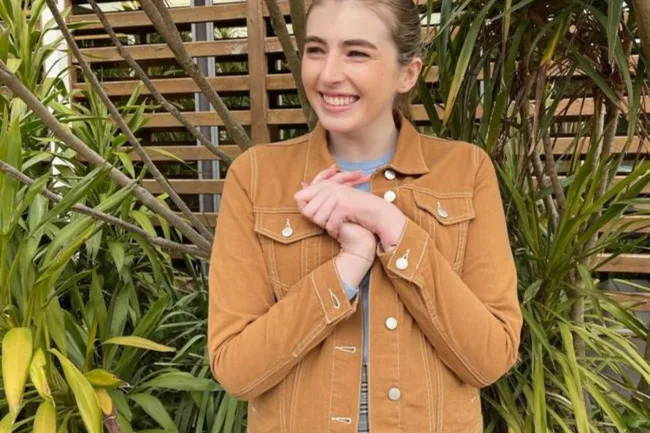 Smiling person in a brown jacket standing in front of a wooden fence and green plants.