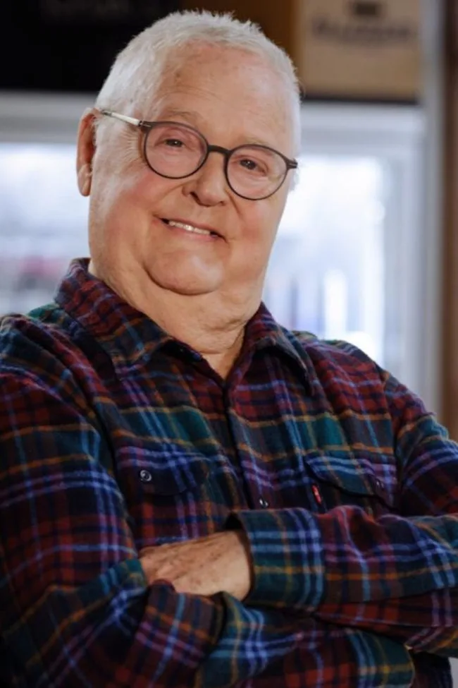 Older man with glasses smiling, wearing a plaid shirt and arms crossed in an indoor setting.