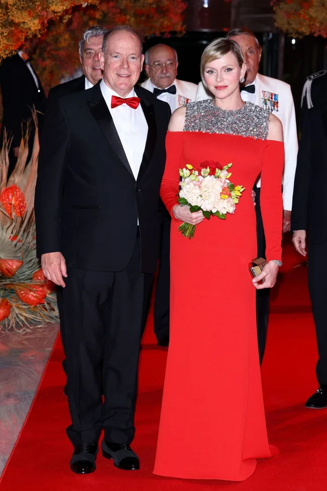 Couple on red carpet; woman in red gown holding bouquet, man in black suit with red bow tie, evening event setting.