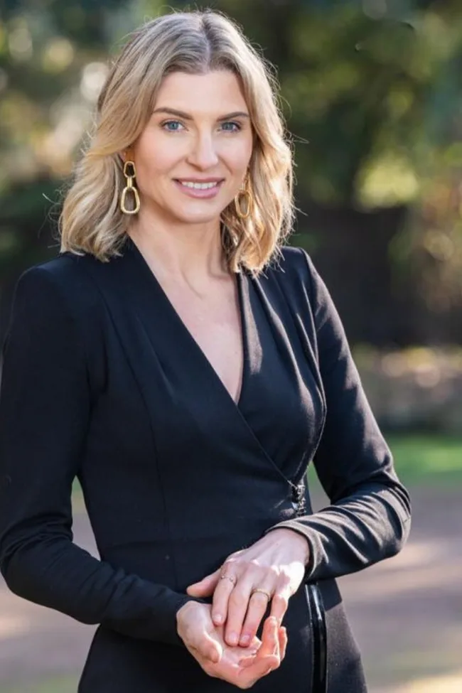 A woman with blonde hair in a black outfit stands outside, smiling with hands clasped.