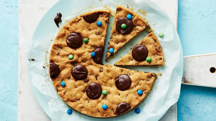 Giant cookie with chocolate and colorful candy toppings, cut into slices, on parchment and blue plate.