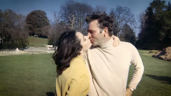 A couple kissing in a park on a sunny day, with trees in the background.