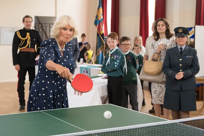 Woman in a polka dot dress playing table tennis, surrounded by people including scouts and military personnel.