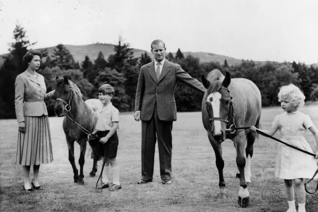 A family in a park with two children holding ponies, surrounded by trees in the background.