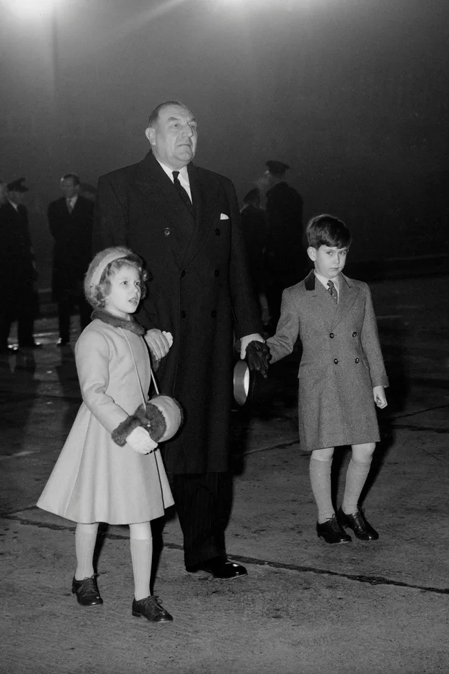 Man in a dark coat with two children holding hands on an airport tarmac at night.