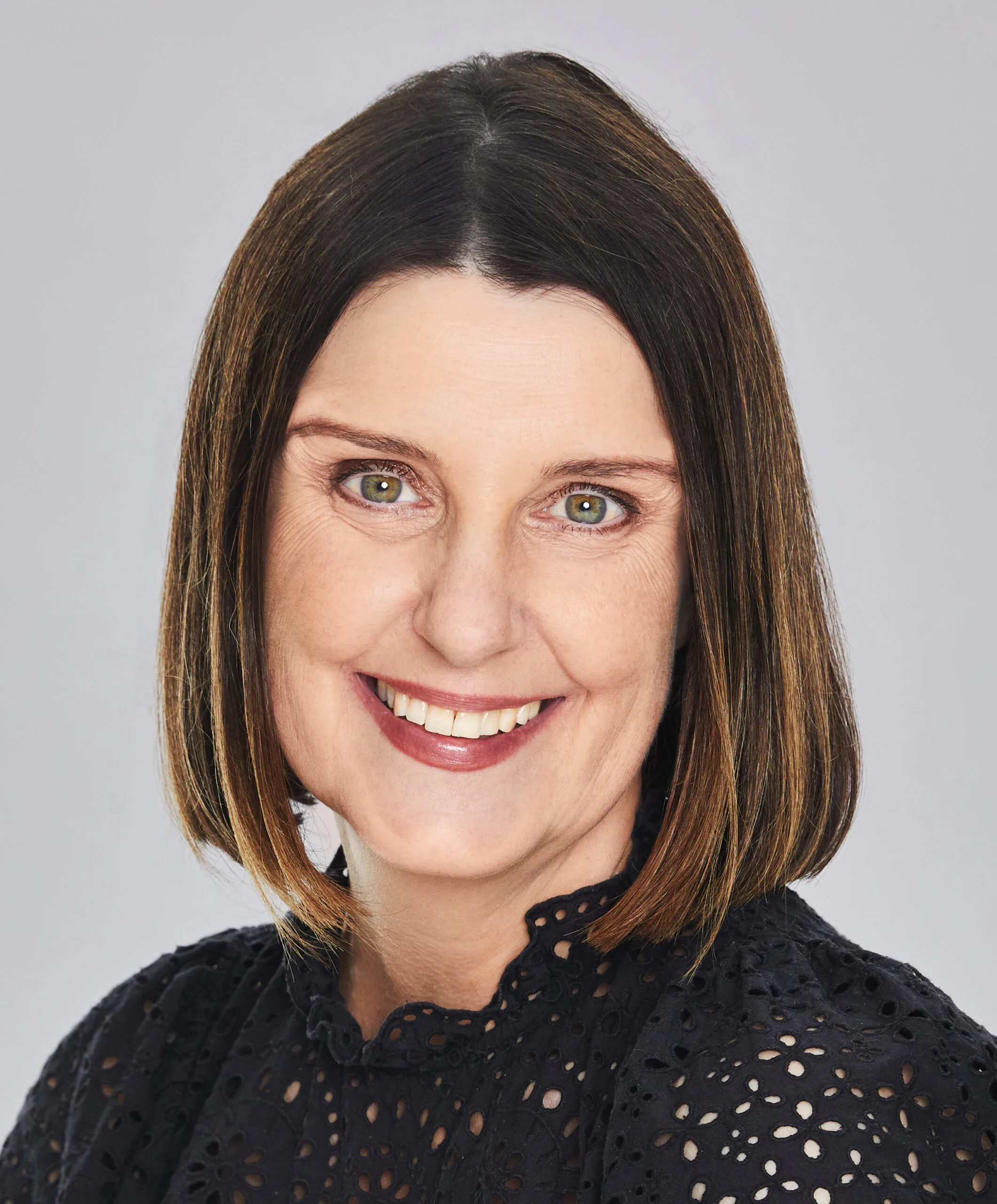 A woman with shoulder-length brown hair and a black lace top smiling at the camera against a gray background.