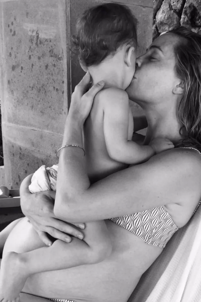 A woman in a bikini embraces and kisses a baby on the cheek in a black and white photo.