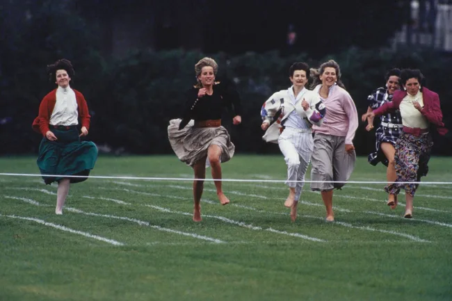 Women in casual attire race barefoot on a grassy field, nearing the finish line with smiles and determination.