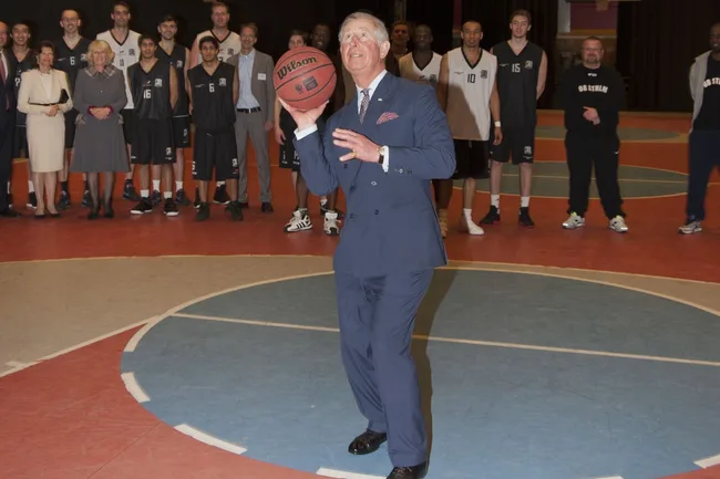 Royal family member in a suit holding a basketball on a court, with a group of people watching in the background.