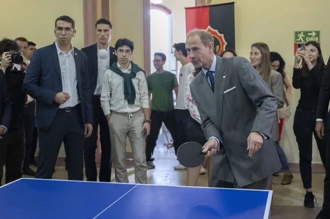 Royal family member playing table tennis indoors, with a small crowd watching attentively.