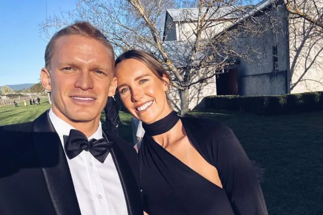 A couple in formal attire smiling outdoors in front of a stone building and trees.