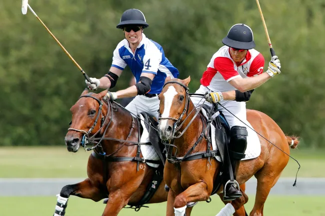 Two men playing polo on horseback, focused and mid-action, wearing helmets and team jerseys on a grassy field.