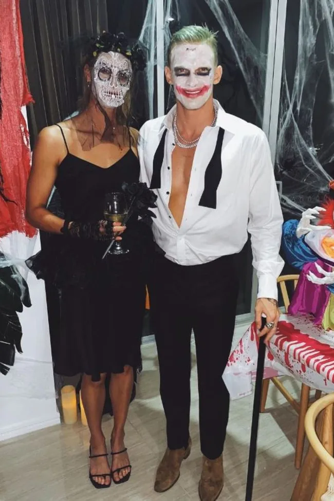 Two people in Halloween costumes; one as a sugar skull, the other resembling the Joker, in a room with spooky decor.
