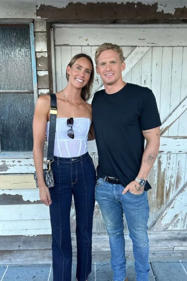 Two people smiling and posing together in casual outfits, standing in front of a rustic wooden door.