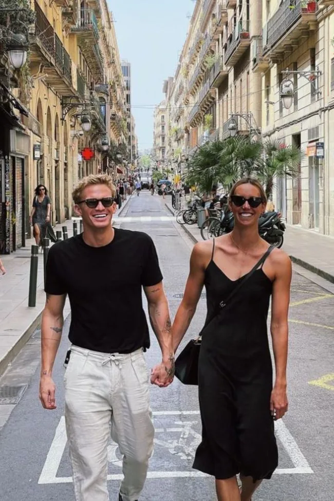 A smiling couple in sunglasses walks hand in hand down a sunny, European-style street lined with historic buildings.
