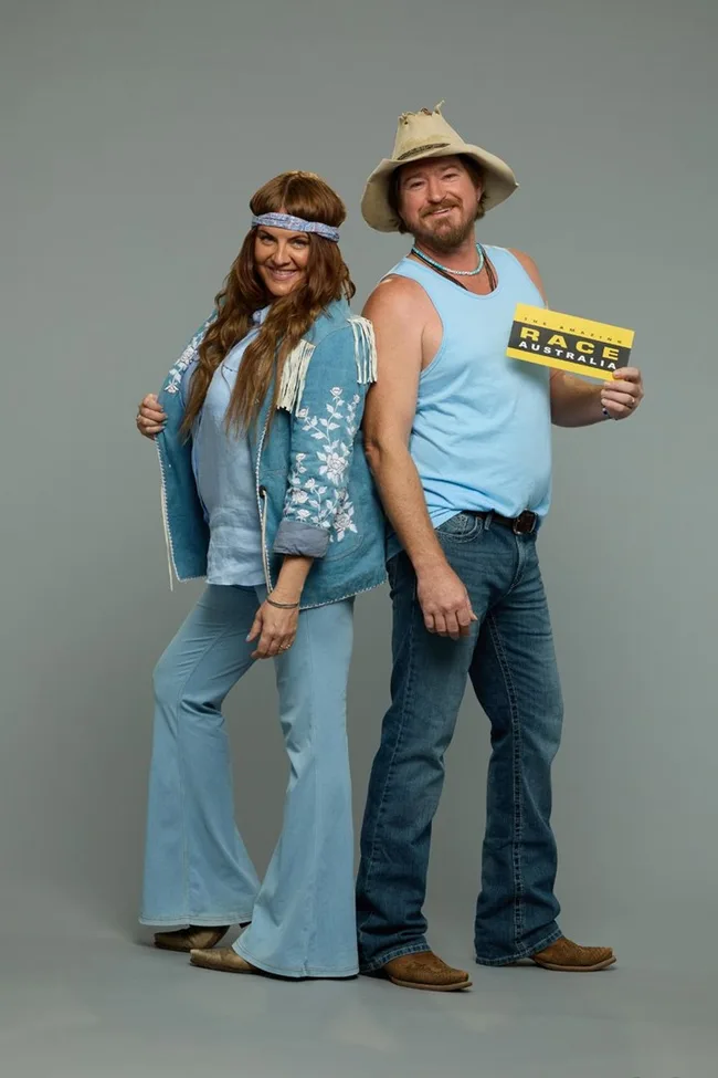 Two people in blue outfits pose cheerfully; one holds an "Amazing Race Australia" card.