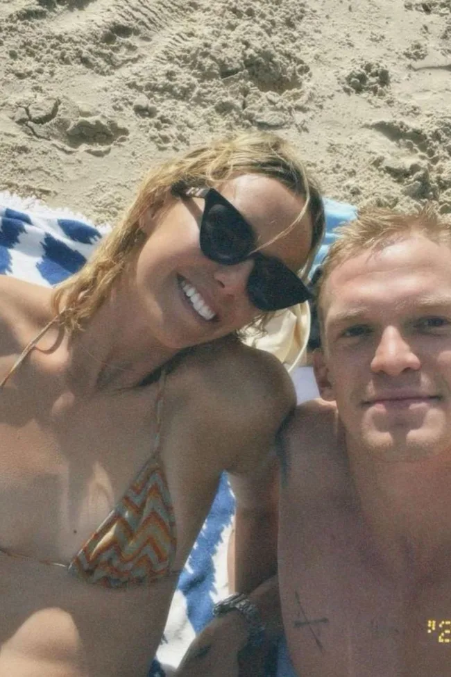 A couple smiles while lying on a beach towel, with sand in the background.
