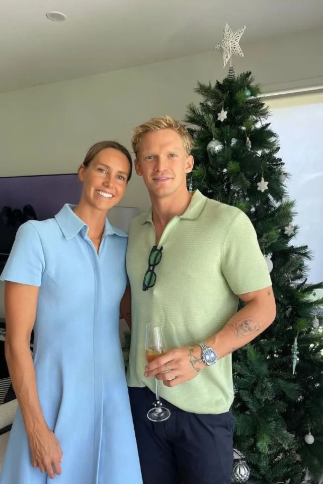 A woman in a blue dress and a man in a green shirt smile beside a decorated Christmas tree indoors.