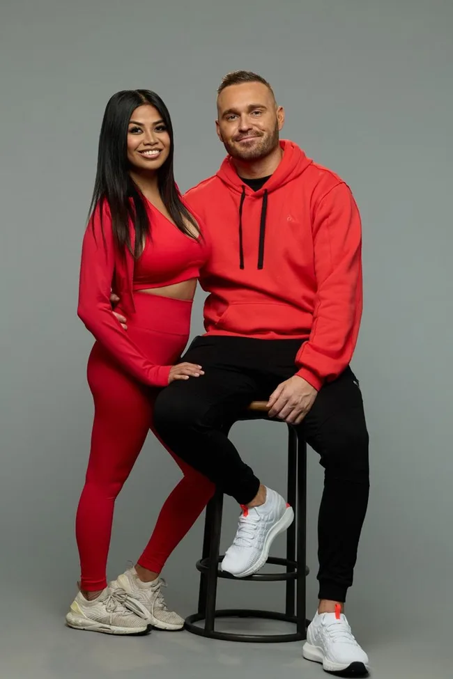 A woman in red and a man in a red hoodie sit closely together; both are smiling at the camera.
