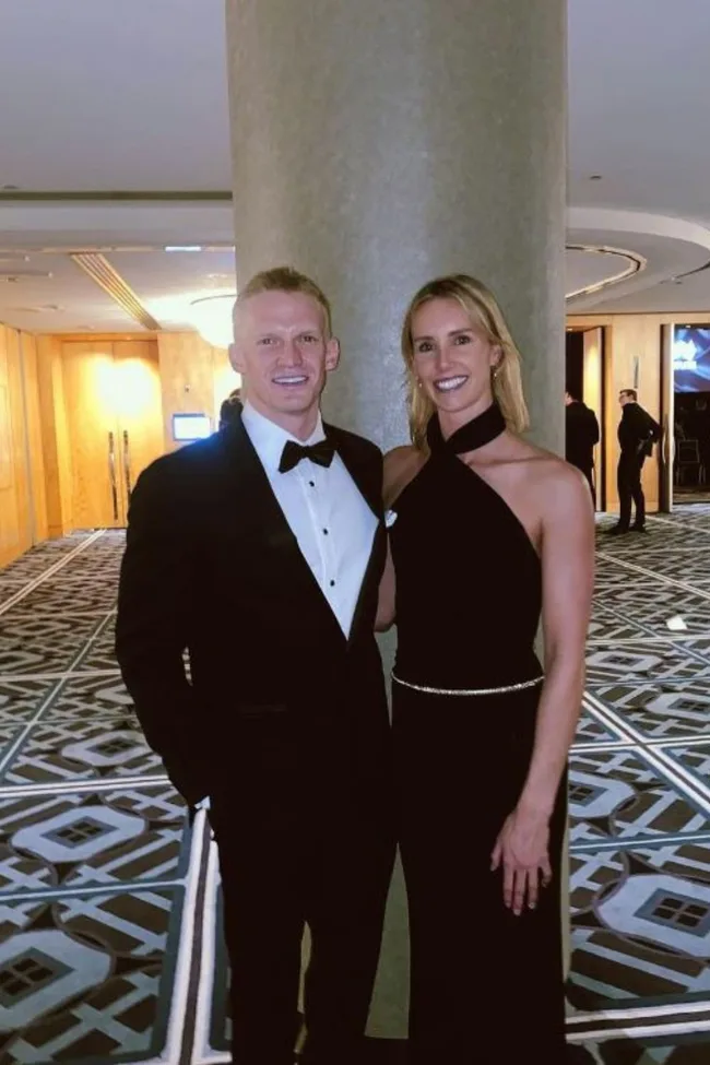 A man in a tuxedo and a woman in a black dress pose together in a hotel hallway.