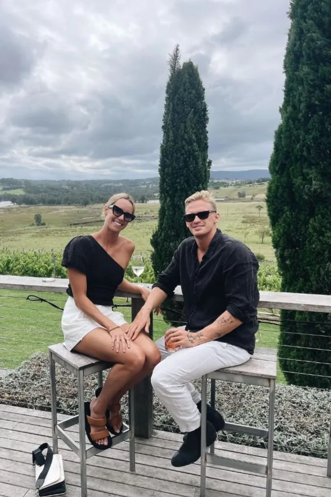 Couple sitting on stools outdoors, smiling, wearing sunglasses; scenic vineyard and trees in the background.