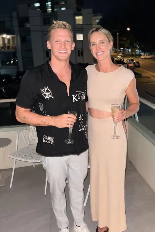 A smiling man in a black shirt and woman in a beige dress holding champagne glasses on a balcony at night.