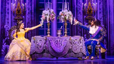 Belle and Beast in ornate clothing toast at a lavishly decorated table in a theatrical production.