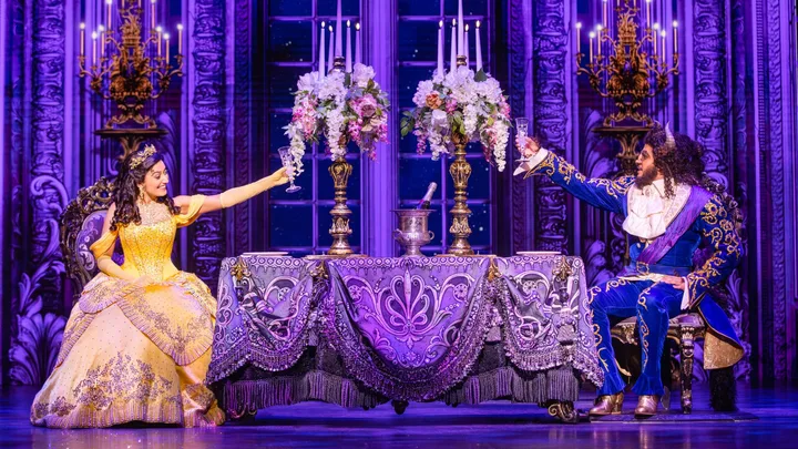 Belle and Beast in ornate clothing toast at a lavishly decorated table in a theatrical production.