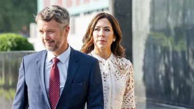 A man in a blue suit and a woman in a white lace dress walking outdoors, looking to the side.
