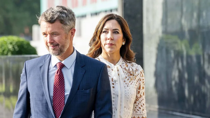 A man in a blue suit and a woman in a white lace dress walking outdoors, looking to the side.