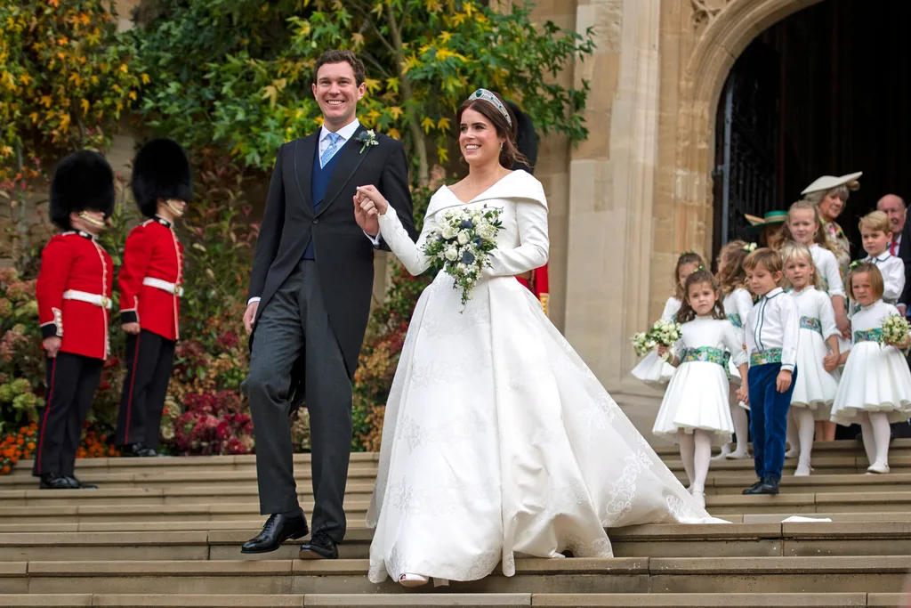 Bride and groom exiting church after wedding, holding hands, surrounded by children and guards in ceremonial attire.