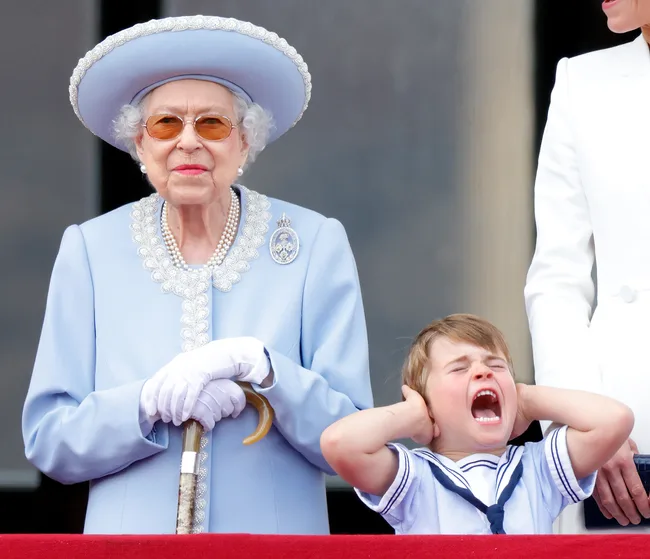 Elderly woman in light blue outfit stands next to a young child covering ears and yelling at a public event.