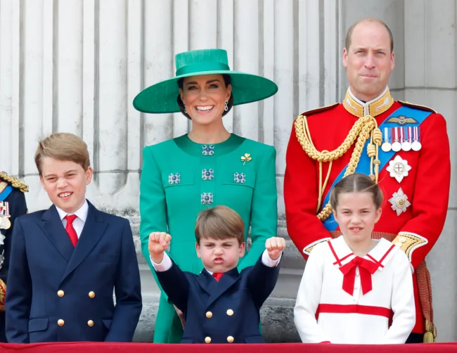 A family of five poses on a balcony, with two adults in formal attire and three children in front, making playful expressions.