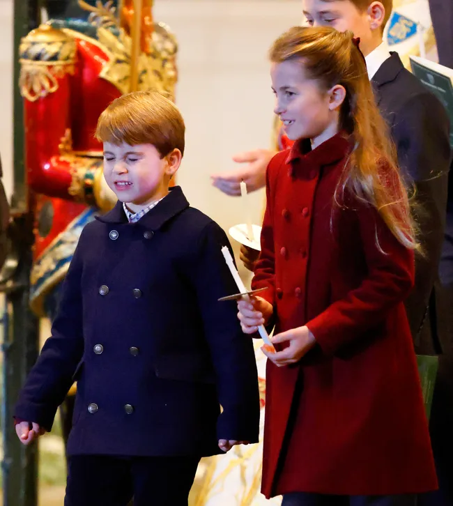 Two children wearing coats holding candles, one squinting and the other smiling, standing indoors with colorful decor behind.