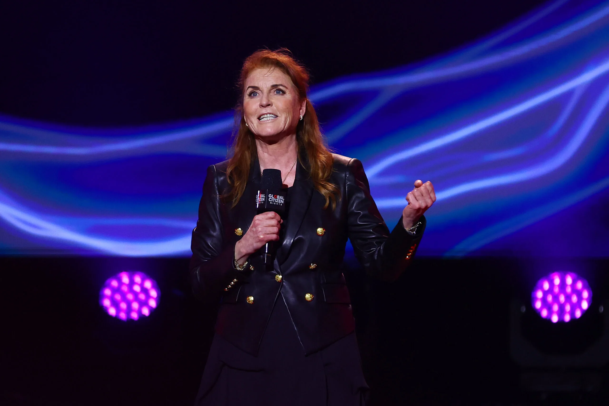 A woman in a black jacket speaks on stage holding a microphone, with a colorful light display in the background.