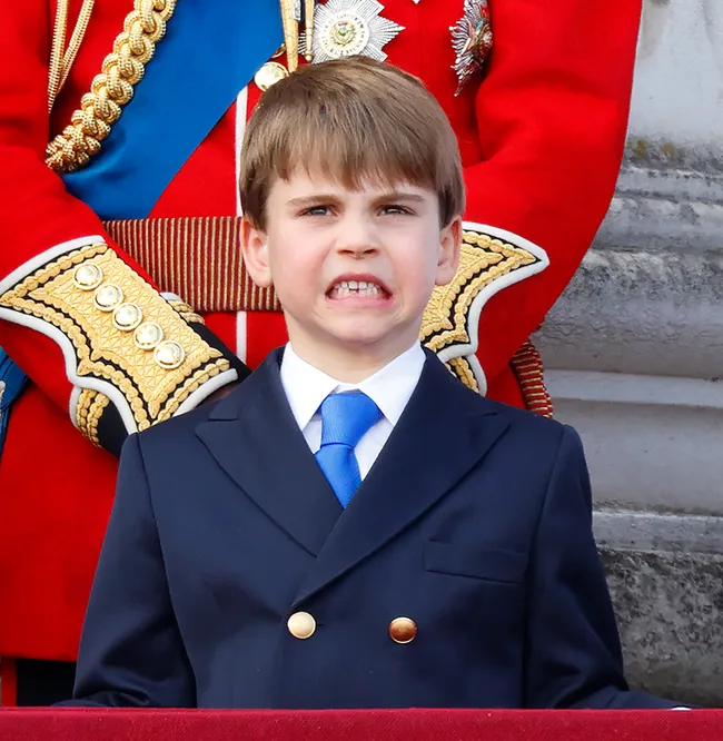 A young boy in a suit grimaces, standing in front of a person in ceremonial uniform with ornate detailing.