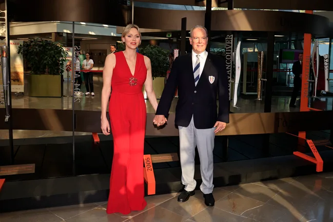 Couple holding hands; woman in red ensemble, man in blazer and tie, standing in a modern indoor setting.