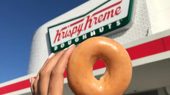 Hand holding a glazed donut in front of a Krispy Kreme store sign under a blue sky.