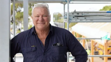 A man in a dark shirt smiling on a construction site, with scaffolding and building materials in the background.