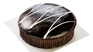 Rich chocolate cake topped with glossy ganache and diagonal white icing lines on a white surface.